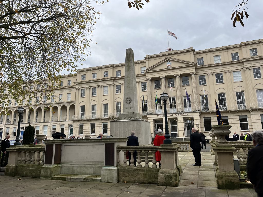 An image of the Cheltenham war memorial statue, shaped like a cone with a square base and head and made of stone, behind a large beige building with the Union Jack on top. The weather is grey and cloudy. There is a tree in the left top corner in front of the statue. There is a group of people stood around the memorial (including the mayor who is wearing a red cloak) and men in smart uniforms.