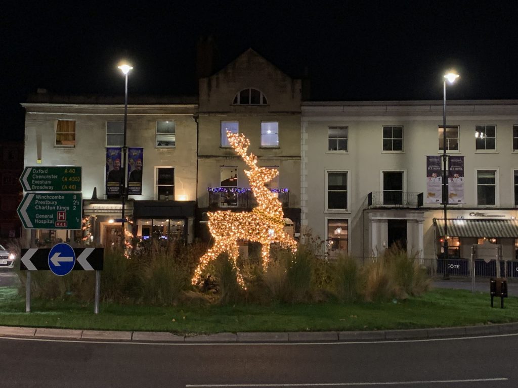 Image Description: photo of a grassy roundabout at night with a golden light reindeer statue, facing the right, in the middle of the roundabout. Behind the roundabout there are three buildings. The building on the left is a restaurant and has cream coloured walls with windows. The building in the middle is taller than the other two and has brown walls. The entrance to the building is blocked by the reindeer. The building on the right is a restaurant, it has cream coloured walls and lots of windows. 