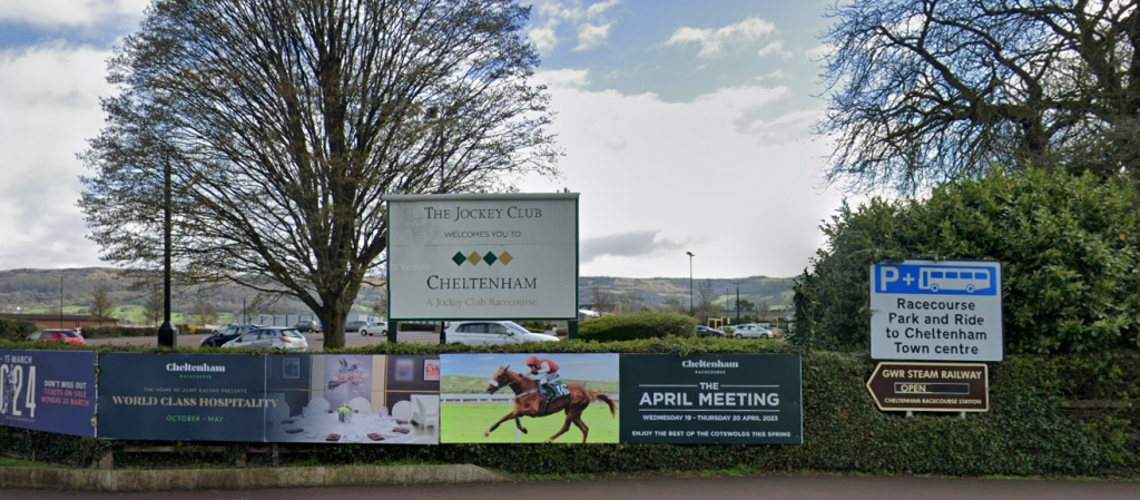 An image of the entrance to Cheltenham racecourse. The sky is blue with clouds. There is a tree in middle behind the white Jockey Club sign and another in the right upper corner. Their leaves are a bit sparse. There are advertisements for the Jockey Club displayed across a long bush just below the entrance sign. There are a few silver cars and a red car in the background in the racecourse carpark.