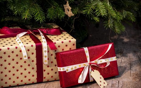 Red foil rectangle present with a white ribbon leaning on a brown paper wrapped box with a red foil ribbon in front of a green Christmas tree