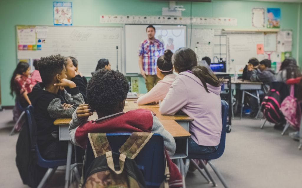 A classroom full of children facing away from the camera towards their teacher and the whiteboard, which is out of focus.